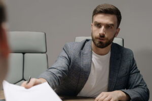Un candidato a un puesto de trabajo, sentado a una mesa de conferencias, entrega documentos a una persona situada en primer plano. Faking en una candidatura: un riesgo innecesario. Antes de recurrir al Faking en una solicitud de empleo, es fundamental considerar las posibles consecuencias.