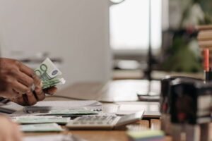 Una mano cuenta billetes de 100 euros sobre un mostrador donde hay varios billetes de euro y una calculadora de sobremesa. Bonos de bienvenida: empresas alemanas buscan trabajadores