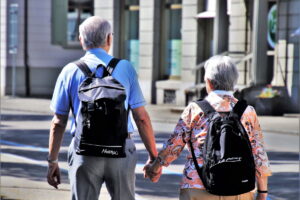 Una pareja de jubilados feliz vista desde atrás, con mochilas - el hombre a la izquierda y la mujer a la derecha -, caminando de la mano por una ciudad. Ingresos de los jubilados en Alemania: conozca los ingresos, el peso de las pensiones, la vivienda y el riesgo de pobreza.