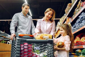 Una familia feliz —madre, padre e hija pequeña— llena su carrito de la compra con alimentos en el supermercado. Reconversión laboral en Alemania: sueldos iniciales, primas y cómo compensar el costo de vida con subsidios oficiales y bonos de contratación.
