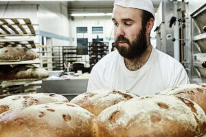 Panadero en su gran panadería ante hogazas de pan recién horneado: A. Producción de Panadería Lutz Panaderos en Alemania exigen reformar la ley para permitir el trabajo dominical de hasta ocho horas y garantizar la producción artesanal.