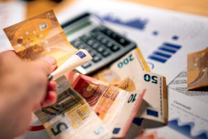 Una mano situada en segundo plano sostiene un billete de euro. Delante de ella hay varios billetes, así como una calculadora de sobremesa y un gráfico impreso. Descubre cómo los beneficios adicionales, la retribución variable y la previsión empresarial transforman los modelos salariales modernos en Alemania.