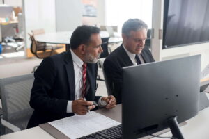Dos empresarios con traje están sentados frente a un ordenador en una oficina moderna y comentan un documento. Las áreas de finanzas, banca y asesoría fiscal lideran la satisfacción laboral en Alemania gracias a su estabilidad y excelentes sueldos.